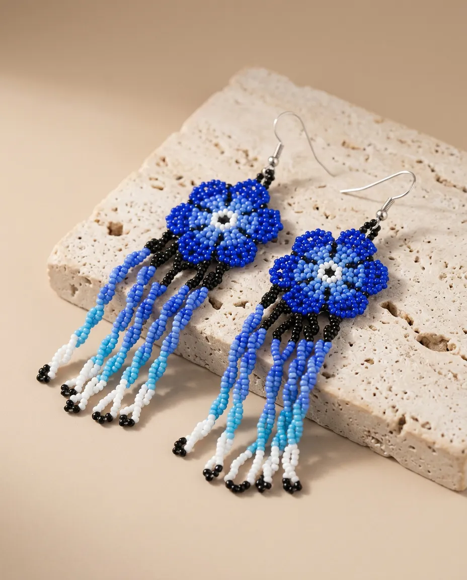 Par de brincos indígenas de missangas em flor azul com franjas longas em degradê do azul ao branco, apoiado sobre uma pedra de travertino clara.