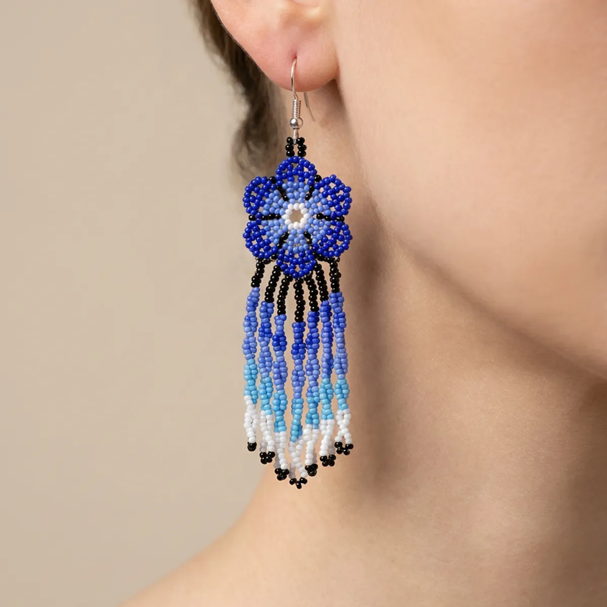Mulher de perfil usando um brinco indígena de missangas em flor azul com franjas longas em degradê do azul ao branco.