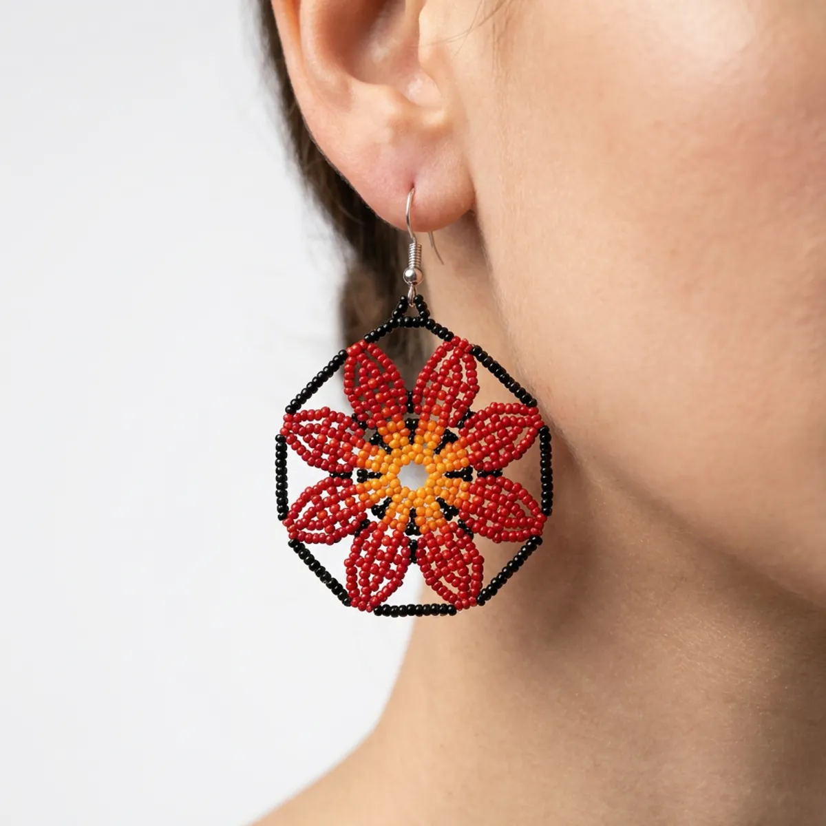 Mulher de perfil usando um brinco indígena de missangas em flor vermelha e laranja com moldura hexagonal preta.