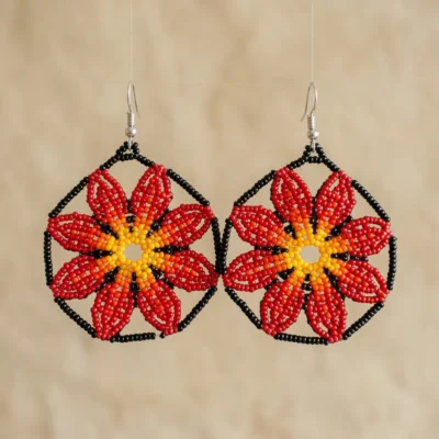 Par de brincos indígenas de missangas em flor vermelha e laranja com moldura hexagonal preta, suspensos lado a lado sobre fundo areia.