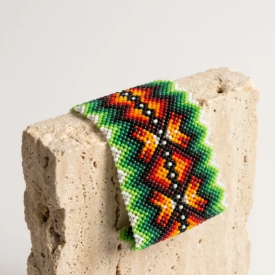 Pulseira indígena de miçanga larga Flechas de Fogo, chevrons em verde, laranja e vermelho sobre base branca, apoiada em bloco de travertino