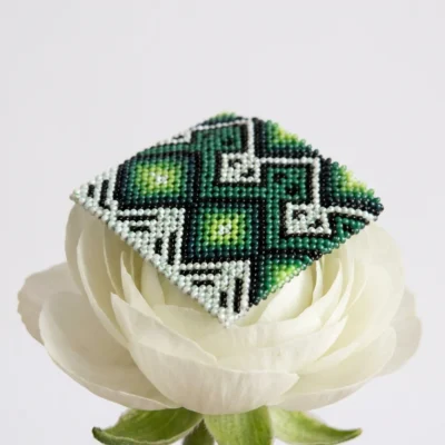 Pulseira indígena de miçanga verde e branca Green Horizont larga, losangos em degradê de verde e turquesa, apoiada sobre flor branca